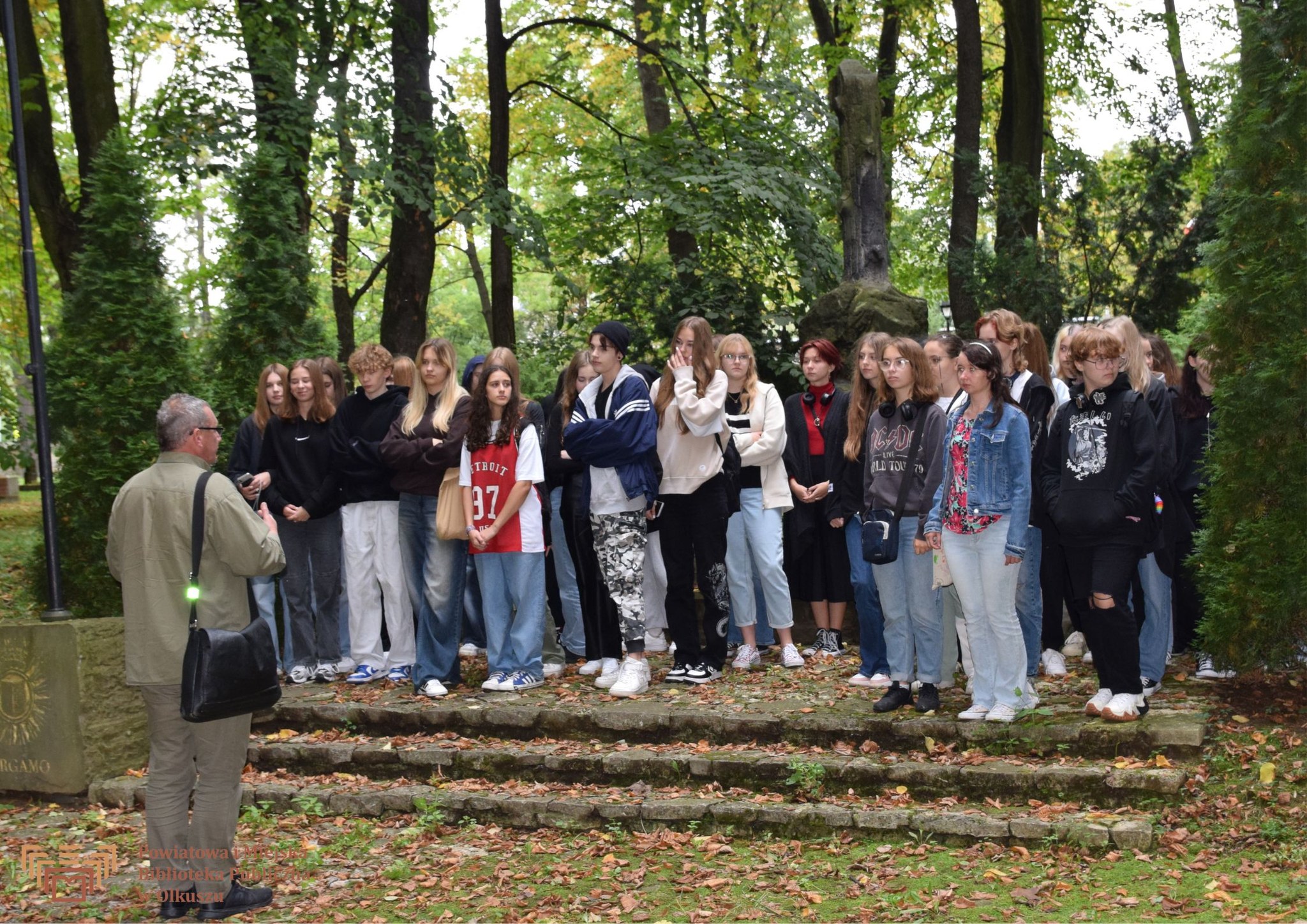 Zdjęcie zostało zrobione na Starym Cmentarzu w Olkuszu. Przedstawia dużą grupę młodzieży. Są zwróceni w stronę mężczyzny, który coś  im opowiada.